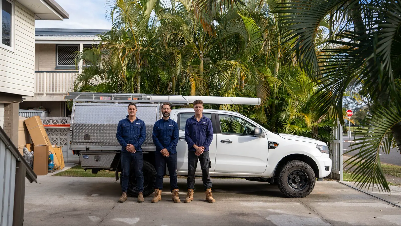 The QMAX crew, three electricians in navy QMAX workwear, standing in front of the white QMAX ute on a Brisbane driveway lined with palms.